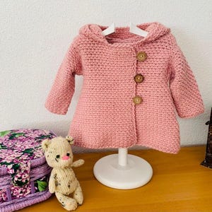 Puede incluir: Un abrigo de ganchillo rosa claro con capucha para bebé o niño pequeño. El abrigo tiene tres botones de madera y se exhibe en un soporte blanco. Un pequeño oso de peluche y una caja decorativa están en primer plano.