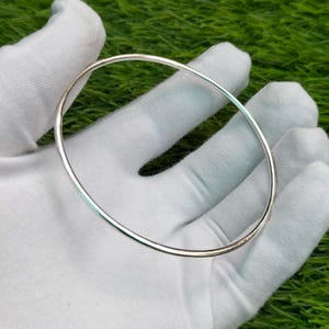 May include: A simple, thin, circular silver bangle bracelet with a smooth, polished, and reflective metallic surface. The bangle features a continuous, unbroken band design with a rounded profile, showcasing a bright, clean, and minimalist aesthetic.
