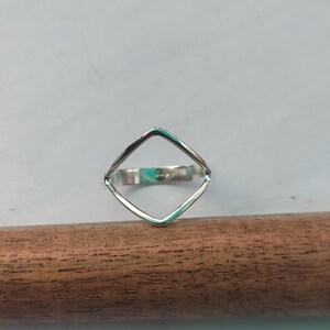 May include: A silver ring with a geometric square design. The ring is set against a light blue background and rests on a wooden dowel. The ring's square shape is open in the center.