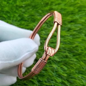 May include: A copper-colored metal bangle bracelet featuring a hook-and-eye style closure mechanism. The design includes a smooth, curved wire loop that secures into a small metal clasp, with decorative coiled wire detailing on both sides of the band.