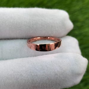 Könnte beinhalten: Ein roségoldener Ring mit gehämmertem Muster. Der Ring wird auf einer weißen Oberfläche vor einem grünen Hintergrund präsentiert. Das Band hat ein flaches Profil und eine leicht unregelmäßige Oberfläche.