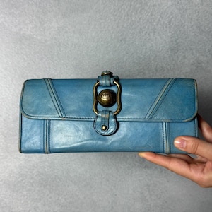 May include: A blue leather wallet with a brass clasp. The wallet is rectangular with stitched detailing and a flap closure. The brass clasp has a decorative design. The wallet is held by a hand.