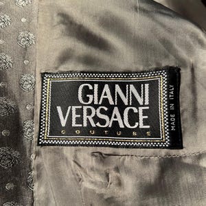 May include: Close-up of the interior of a jacket, featuring a label that reads "GIANNI VERSACE COUTURE" in white text on a black background, framed with gold and white trim. The lining is a muted gray-green color, with a subtle pattern.