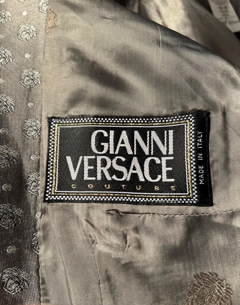 May include: Close-up of the interior of a jacket, featuring a label that reads "GIANNI VERSACE COUTURE" in white text on a black background, framed with gold and white trim. The lining is a muted gray-green color, with a subtle pattern.