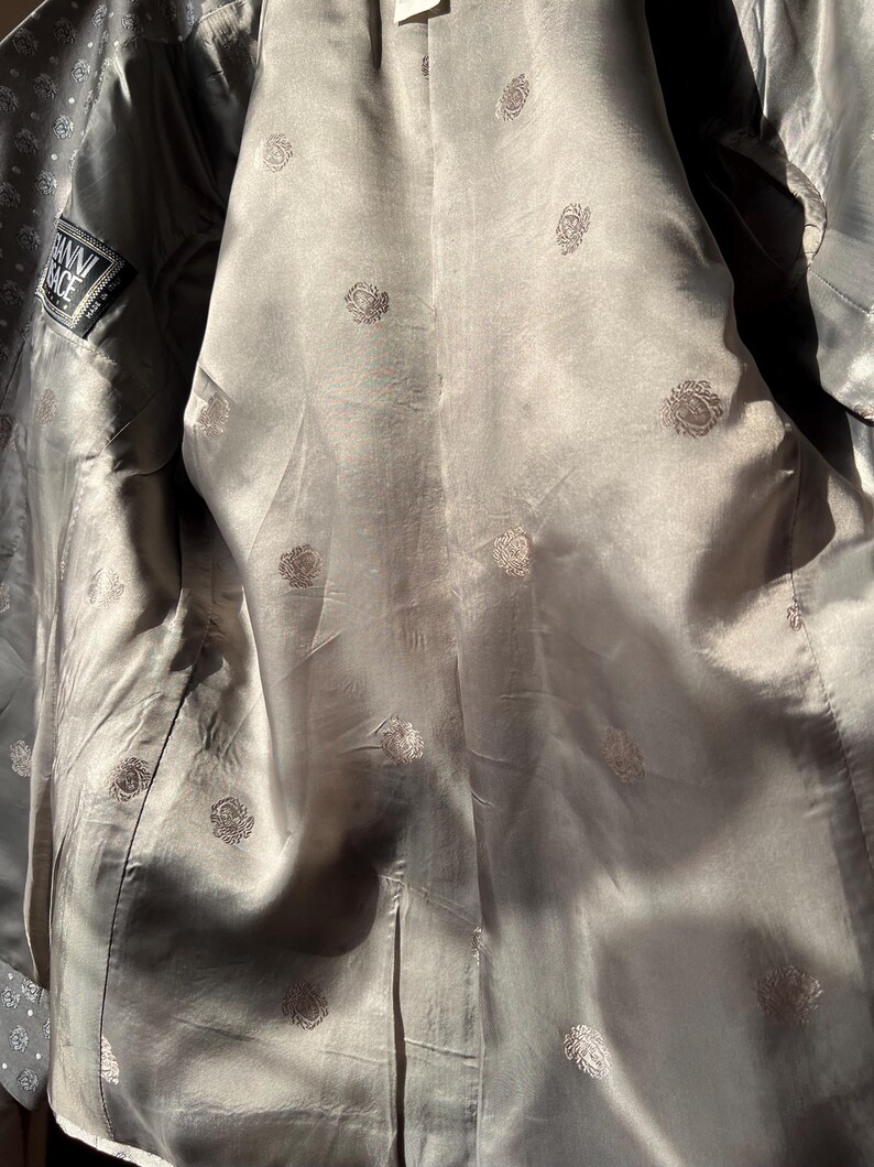 May include: Interior view of a gray blazer with a silky lining. The lining is patterned with small, repeating floral designs. A label with the text "Gianni Versace" is visible on the inside.