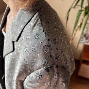 May include: A gray blazer with a floral pattern and white polka dots. The blazer has a notched lapel and a tailored fit. The fabric appears to be a lightweight material, suitable for warmer weather. The floral design is a repeating pattern of stylized roses.