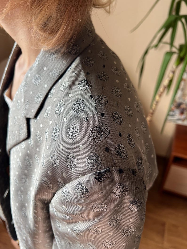 May include: A gray blazer with a floral pattern and white polka dots. The blazer has a notched lapel and a tailored fit. The fabric appears to be a lightweight material, suitable for warmer weather. The floral design is a repeating pattern of stylized roses.