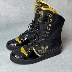 May include: Black and gold high-top sneakers with a glossy finish. The sneakers feature gold accents, including the iconic three-stripe logo and eyelets. The tongue area has text and a crown logo.