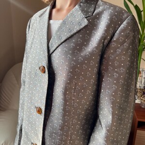 May include: A gray blazer with a repeating floral pattern and white polka dots. The blazer has three brown buttons and a notched lapel. The blazer is a classic style.