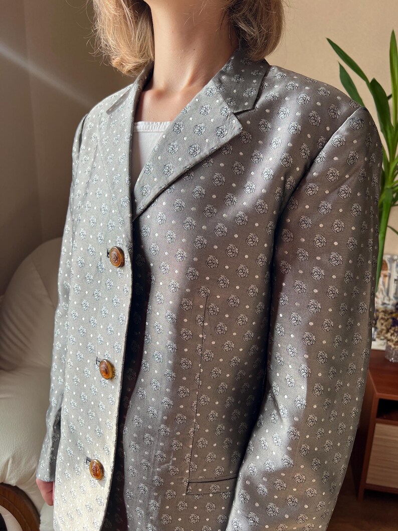 May include: A gray blazer with a repeating floral pattern and white polka dots. The blazer has three brown buttons and a notched lapel. The blazer is a classic style.
