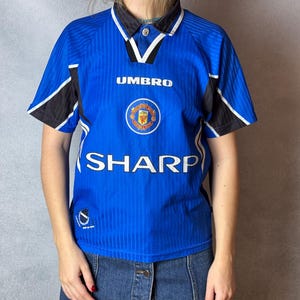 Puede incluir: Camiseta de fútbol azul con detalles en negro y blanco. La camiseta presenta el logo de Umbro, un escudo del equipo y el logo de Sharp. Tiene cuello y mangas cortas. La camiseta se lleva con una falda vaquera.