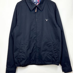 May include: A black jacket with a collared neckline and a zip fastening. The jacket has two front pockets and a small logo on the chest. The interior collar lining features a plaid pattern. The jacket is hanging on a white hanger.