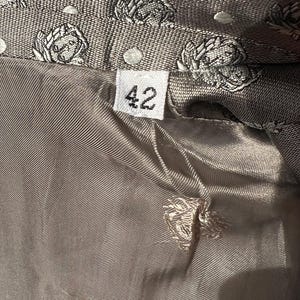 May include: Close-up of a gray jacket lining with a floral pattern and the number "42" on a tag. The jacket's exterior features a repeating pattern of white floral designs on a textured gray fabric. The lining is a smooth, solid gray.