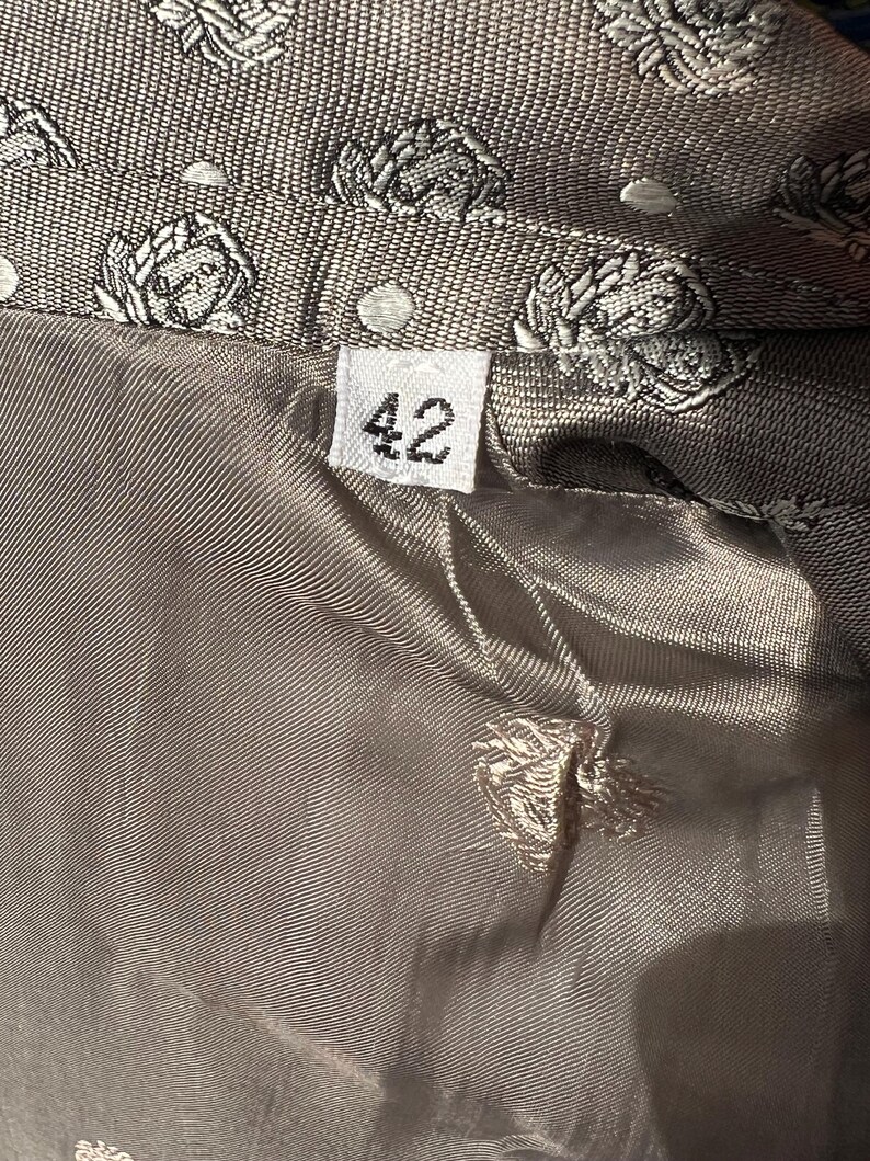 May include: Close-up of a gray jacket lining with a floral pattern and the number "42" on a tag. The jacket's exterior features a repeating pattern of white floral designs on a textured gray fabric. The lining is a smooth, solid gray.