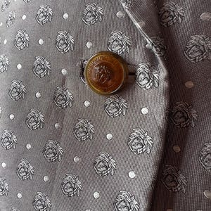 May include: Close-up of a gray blazer with a repeating pattern of floral crests and small white dots. The blazer has a large, round, amber-colored button with a detailed emblem. The fabric has a subtle sheen.