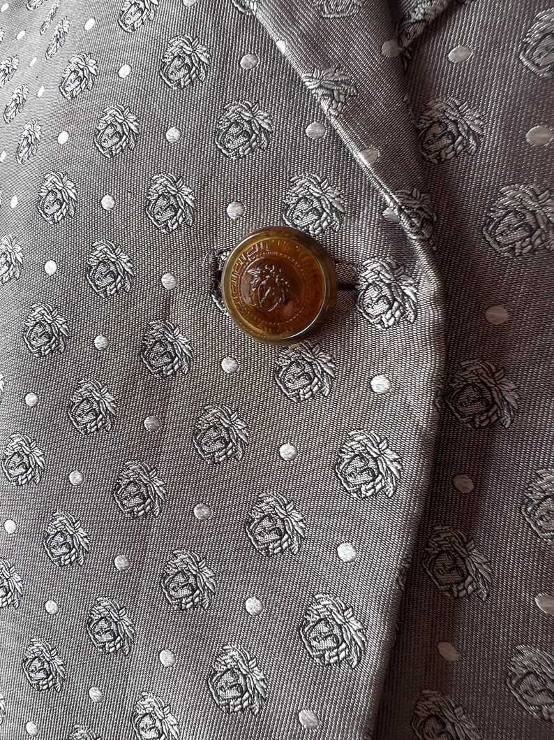 May include: Close-up of a gray blazer with a repeating pattern of floral crests and small white dots. The blazer has a large, round, amber-colored button with a detailed emblem. The fabric has a subtle sheen.