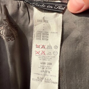 May include: Close-up of a label from a garment, with text in multiple languages. The label is white with black text, and the garment is a patterned gray fabric. The label indicates the garment is made in Italy and lists fabric content and care instructions.