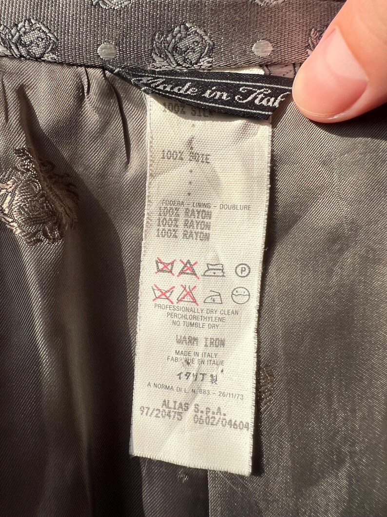 May include: Close-up of a label from a garment, with text in multiple languages. The label is white with black text, and the garment is a patterned gray fabric. The label indicates the garment is made in Italy and lists fabric content and care instructions.