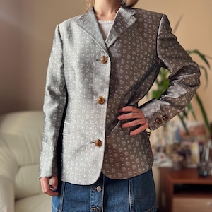 May include: A silver blazer with a subtle floral pattern and three brown buttons. The blazer has long sleeves and a notched collar. The fabric has a slight sheen. The blazer is paired with a denim skirt.