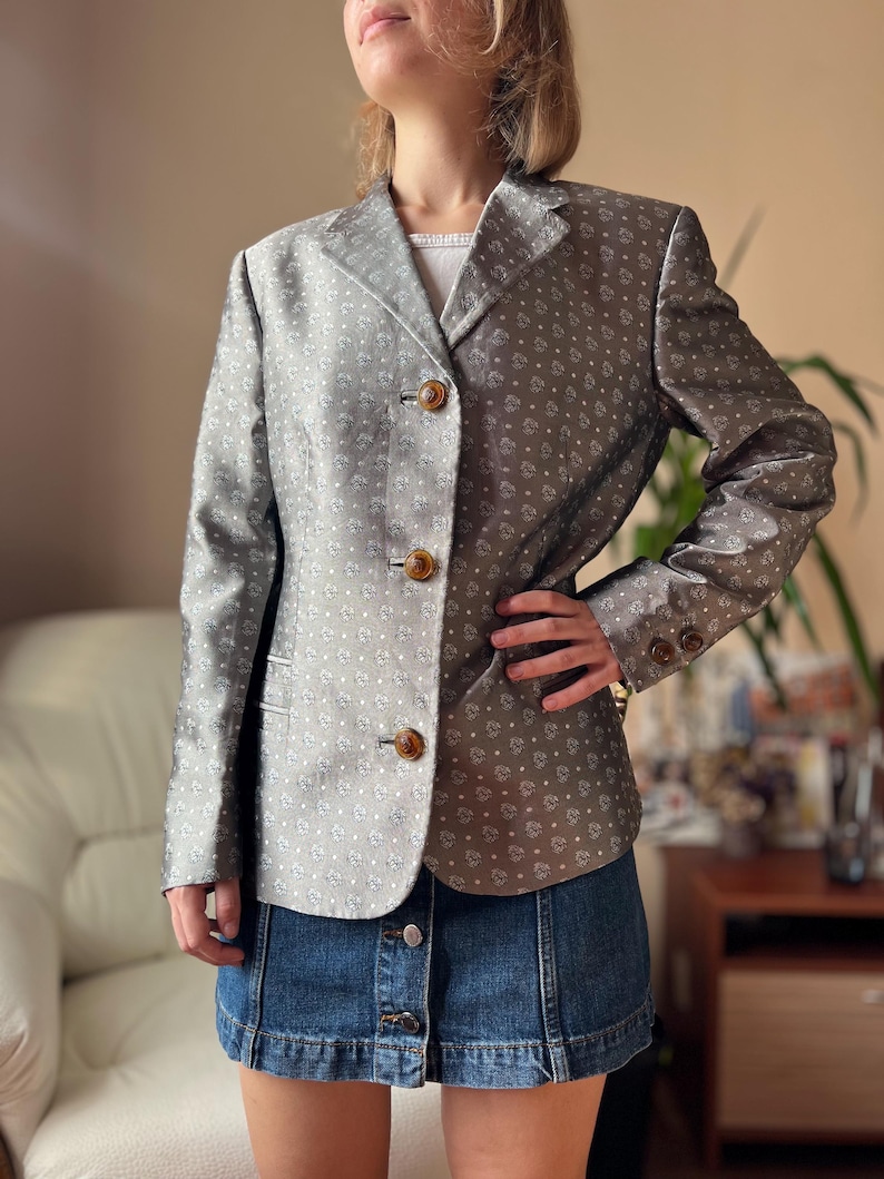 May include: A silver blazer with a subtle floral pattern and three brown buttons. The blazer has long sleeves and a notched collar. The fabric has a slight sheen. The blazer is paired with a denim skirt.