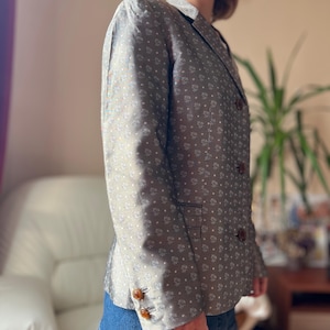 May include: A gray blazer with a white floral pattern and brown buttons. The blazer has a classic design with a notched collar and a double-breasted closure. The sleeves have three buttons. The blazer is paired with a denim skirt.