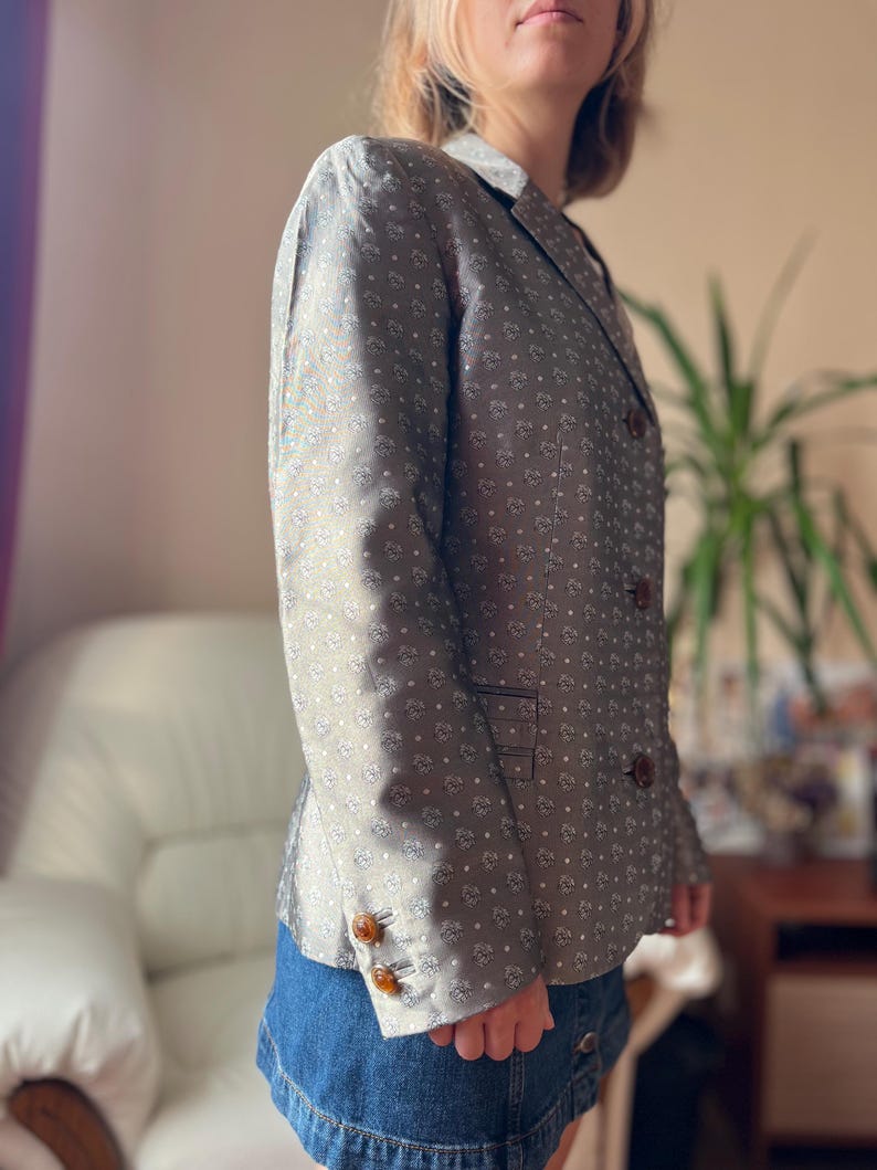 May include: A gray blazer with a white floral pattern and brown buttons. The blazer has a classic design with a notched collar and a double-breasted closure. The sleeves have three buttons. The blazer is paired with a denim skirt.
