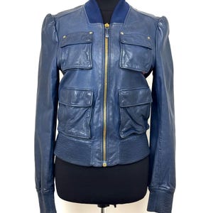 May include: A navy blue leather bomber jacket with a gold zipper and four front pockets. The jacket has a ribbed collar, cuffs, and hem. Small gold stars are on the shoulders. The jacket is displayed on a black mannequin.