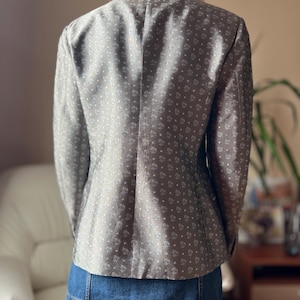 May include: A gray blazer with a subtle floral pattern and a classic tailored fit. The jacket is worn over a denim skirt. The blazer's fabric has a soft sheen, and the design is a repeating pattern of small, stylized flowers.