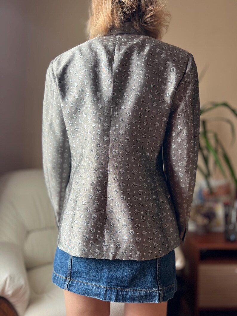 May include: A gray blazer with a subtle floral pattern and a classic tailored fit. The jacket is worn over a denim skirt. The blazer's fabric has a soft sheen, and the design is a repeating pattern of small, stylized flowers.