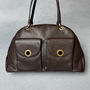 May include: A dark brown leather handbag with two front pockets, each featuring a gold-coloured circular emblem. The bag has a curved top and two slender handles, with gold-coloured hardware at the handle attachments.