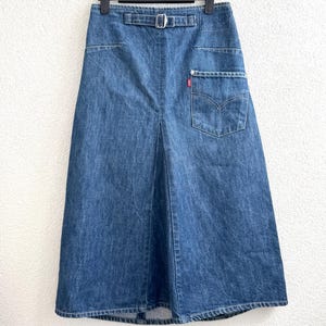 May include: A denim skirt in a medium wash, featuring a belt with a silver buckle, front pockets, and a back pocket with a red Levi's tag. The skirt is hanging on a black hanger.