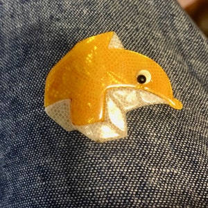 Puede incluir: Broche con forma de delfín amarillo con un patrón texturizado en forma de escamas. El broche tiene una parte inferior blanca y un pequeño ojo negro. El delfín está sobre un fondo de mezclilla azul.