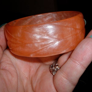 Op de afbeelding: Een doorschijnende oranje armband met een gesneden bladerenpatroon. De armband wordt in een hand gehouden, waardoor de kleur en textuur worden benadrukt. Dit sieraad is een stijlvol accessoire.