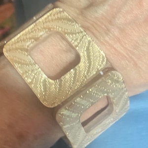 Könnte beinhalten: Ein goldfarbenes Armband mit zwei großen, quadratischen Gliedern. Jedes Glied hat eine strukturierte, gemusterte Oberfläche und einen quadratischen Ausschnitt in der Mitte. Das Armband wird am Handgelenk getragen.