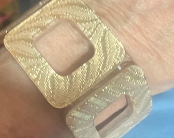 Puede incluir: Una pulsera dorada con dos eslabones grandes y cuadrados. Cada eslabón tiene una superficie texturizada y estampada, con un recorte cuadrado en el centro. La pulsera se lleva en la muñeca.