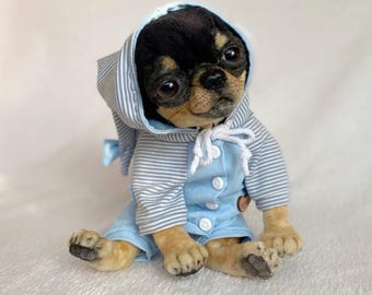 May include: A plush Chihuahua puppy wearing a light blue and white striped hooded jumpsuit. The puppy has black and tan fur and is sitting on a white surface. The jumpsuit has white buttons and a small wooden button.