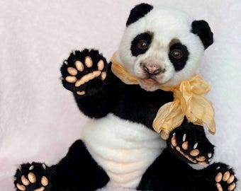 May include: A plush panda bear with black and white fur, sitting with its paws outstretched. The panda has a white face with black patches around the eyes and ears. A yellow ribbon is tied around its neck. The bear's paws have tan-colored pads.