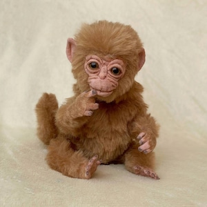 May include: A plush, brown monkey doll with a thoughtful expression. The toy monkey has a light brown face, ears, and hands, with a darker brown fur body. The monkey is sitting and appears to be touching its chin.