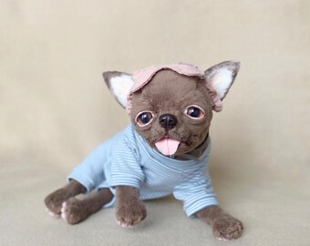 May include: A brown Chihuahua plush toy wearing a light blue striped shirt and a pink cap. The dog has its tongue sticking out and is lying down. The toy has large, expressive eyes and pink paws.