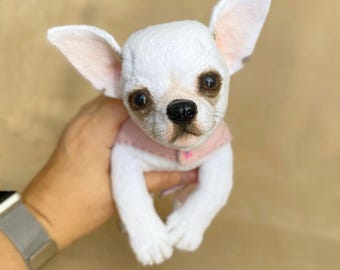 May include: A white Chihuahua dog figurine with large, upright ears and dark eyes. The dog is wearing a light pink sweater. The figurine is held in a person's hand.