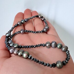 Könnte beinhalten: Halskette mit dunkelgrauen Perlen und kleinen, facettierten schwarzen Perlen. Die Perlen haben einen glänzenden Glanz und schaffen ein elegantes Accessoire. Die Halskette wird in einer Hand gehalten.