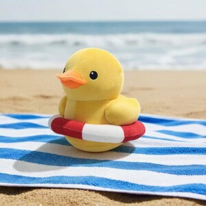 Peut inclure: Un canard en peluche jaune avec un bec orange, portant une bouée de sauvetage rouge et blanche, posé sur une serviette de plage à rayures bleues et blanches. La plage de sable et la mer sont en arrière-plan.