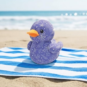 May include: A soft purple duck toy with an orange beak and black eyes, patterned with white seashells and seaweed. The duck sits on a blue and white striped towel on a sandy beach, with the sea visible in the background.