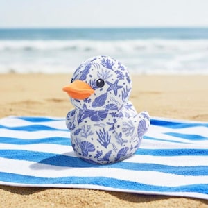 May include: A soft duck toy featuring a white base with a blue seashell design. The duck has an orange beak and black eyes. It rests on a blue and white striped towel on a sandy beach, with the sea visible in the background.