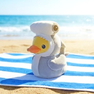 May include: A plush yellow duck wearing a shark costume and a white hat, sitting on a blue and white striped towel on a sandy beach. The duck has an orange beak and black eyes. The background shows the ocean and blue sky.