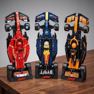Könnte beinhalten: Drei Formel-1-Rennwagenmodelle in Rot, Blau und Orange. Jedes Auto steht auf einer schwarzen Basis mit Sponsorenlogos und dem F1-Logo. Die Autos haben detaillierte Designs, einschließlich Rädern, Flügeln und Fahrerfiguren. Die Autos werden auf einer Holzoberfläche präsentiert.