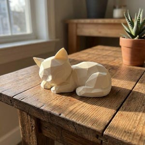 Peut inclure: Une sculpture de chat géométrique de couleur crème, posée sur une table en bois rustique. Le chat est dans une pose détendue, les yeux fermés. La table est en bois vieilli, et l'arrière-plan comprend une fenêtre et une plante en pot.