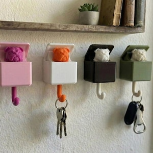 May include: Four wall-mounted key holders in pink, white, black, and olive green. Each has a bulldog head inside a box with a hook below. Keys hang from two of the holders. A shelf with books and a plant is in the background.