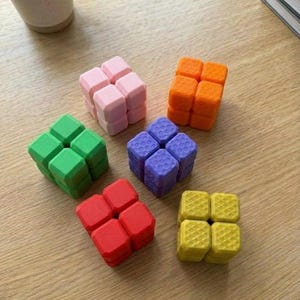 May include: A collection of colorful fidget cubes in shades of pink, orange, green, purple, red, and yellow. Each cube is composed of smaller, textured blocks. The cubes are arranged on a light wooden surface, with a mug and books in the background.