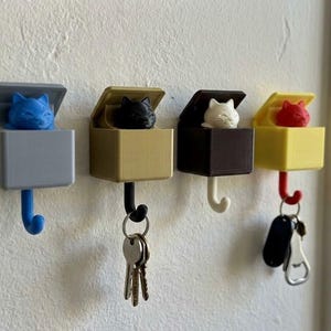Puede incluir: Cuatro llaveros con forma de gato en fila, cada uno con un gato y una caja de diferente color. Los colores son azul, negro, blanco y rojo. Cada soporte tiene un gancho debajo para llaves u otros objetos pequeños. Los soportes están montados en una pared blanca.